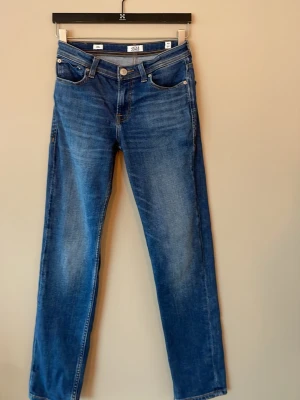 Blå raka jeans från Jack & Jones - Säljer ett par klassiska blå jeans från Jack & Jones med rak passform och fem fickor. Jeansen har snygga slitningar framtill och en patch med logga bak i midjan. Tillverkade i denim med kontrastsömmar och normal midja.