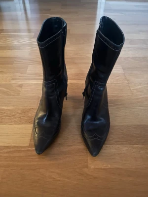 ECCO högklackade skinnstövletter – svart –storlek 38 - Eleganta högklackade stövletter från ECCO i svart med spetsig tå och fina brogue-detaljer. Dragkedja på insidan gör dem lätta att ta på och av, och klacken ger både stil och komfort. Skorna är i bra skick och perfekta för både vardag och fest. Storleken är 38. Skicka bara meddelande ifall ni undrar något mer