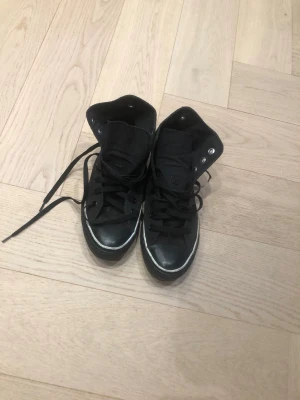 Svarta höga sneakers i canvas - Säljer ett par svarta höga sneakers med klassisk design och vita detaljer på sulan. Skorna har svart canvasmaterial, svarta skosnören och metallhål för snörningen. Perfekta för en avslappnad streetstil.