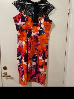 Blommig klänning med spetsdetalj - Säljer en färgstark blommig klänning med orange, rosa, marinblå och vit som dominerande färger. Klänningen har en snygg svart spets upptill och knyt i nacken. Modellen är figurnära och knälång, perfekt för dig som gillar att sticka ut med mönster och detaljer.