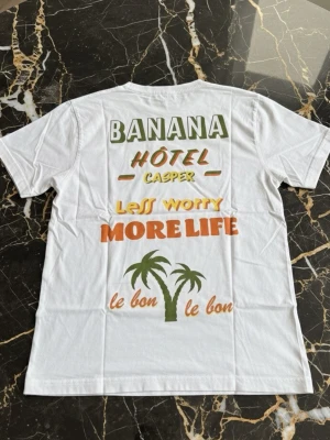 Vit t-shirt med färgglad text och palmer - Vit t-shirt med tryck i grönt, gult, orange och brunt. Texten på ryggen har ord som 'BANANA', 'HÔTEL', 'LESS WORRY MORE LIFE' och två palmer. Avslappnad passform och klassisk rund hals. Perfekt för en chill och färgglad stil.