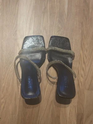 Svarta sandaler med strass från Simmi - Säljer ett par svarta sandaler från Simmi med fyrkantig tå och tunna remmar dekorerade med glittrande strass. Sandalerna har en elegant design och låg klack, perfekta för att lyfta vilken outfit som helst. Materialet är konstläder med detaljer i metall och strass.
