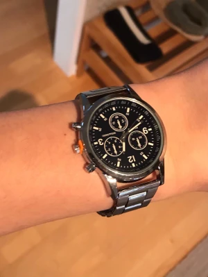 Högkvalitets klocka - Snygg kronografklocka med svart urtavla och tre mindre subdials. Klockan har tydliga vita siffror och markeringar samt en robust länk i blankt silverfärgat metall. Perfekt accessoar för dig som gillar en klassisk men sportig look.