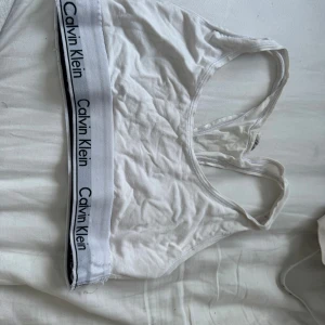 Vit sport-bh från Calvin Klein - Vit sport-bh från Calvin Klein med bred resår och logga runt om. Bh:n har brottarrygg och är tillverkad i mjukt bomullsmaterial för extra komfort. Perfekt för en avslappnad och sportig stil.