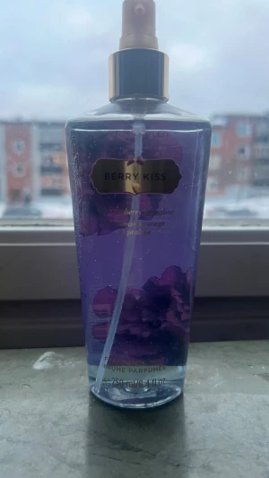 Victoria's Secret Berry Kiss Body Mist - En lila body mist från Victoria's Secret med doften Berry Kiss. Flaskan är genomskinlig med guldigt spraymunstycke och har en volym på 250 ml. Doften har inslag av bär och viol, och flaskan har en elegant, feminin design. Använd en gång 