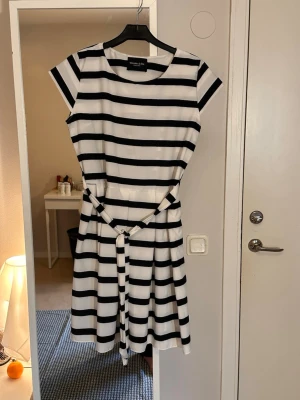 Randig kortklänning med knytband - Säljer en svartvit randig kortklänning från WERA STOCKHOLM. Klänningen har rund hals, korta ärmar och ett matchande knytband i midjan för en snygg siluett. Perfekt för en fräsch och stilren look.
