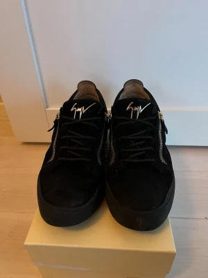 Svarta sneakers från Giuseppe Zanotti - Säljer ett par svarta sneakers från Giuseppe Zanotti med snygga silverfärgade dragkedjor på sidorna och ikonisk logga på plösen. Skorna är låga och har ovandel i mocka samt svarta snören. Perfekta för dig som gillar stilrena och exklusiva detaljer.