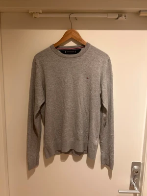 Grå tröja från Tommy Hilfiger - Klassisk grå tröja från Tommy Hilfiger i mjuk bomullsblandning med rund halsringning. Liten broderad logga på bröstet och diskreta ribbade muddar vid ärmslut och nederkant. Perfekt basplagg för en clean och stilren look.
