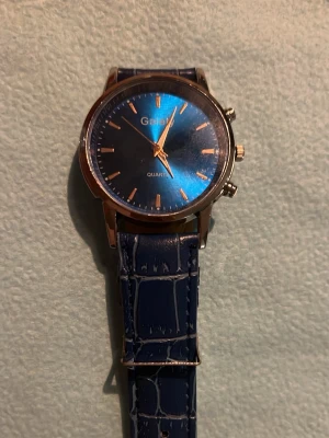 Blå klocka med läderband - Stilren klocka från Galex med blå urtavla och guldfärgade detaljer. Armbandet är i blått konstläder med krokodilpräglat mönster. Klockan har tre knappar på sidan och klassisk analog visning. Perfekt accessoar för att lyfta din outfit.