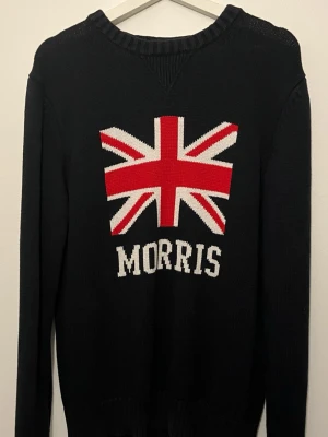 Svart stickad tröja Morris - Stickad svart tröja från Morris med Union Jack-flagga i rött, vitt och blått på bröstet samt Morris-logga under. Tröjan har rund halsringning och långa ärmar. Perfekt för dig som gillar klassisk brittisk stil och vill ha något som sticker ut.