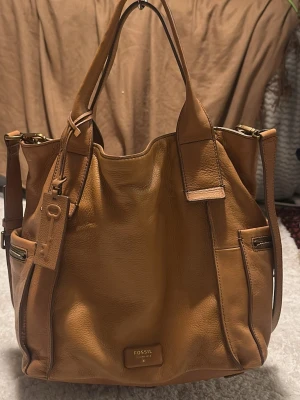 Kamelbrun handväska från Fossil - Snygg kamelbrun handväska från Fossil i mjukt läder. Väskan har dubbla handtag, avtagbar axelrem och flera ytterfack med dragkedja. Dekorativ nyckelbricka och Fossil-logga i guld framtill. Perfekt storlek för allt du behöver ha med dig.