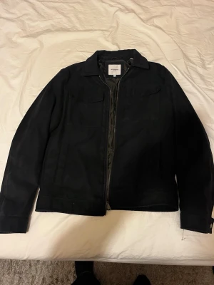 Svart overshirt från jack and jones - Stilren svart overshirt från jack and jones med dragkedja framtill och två bröstfickor. Overshirten har klassisk krage och är helfodrad med slätt tyg på insidan. Perfekt för lager-på-lager och enkel att matcha med olika outfits.