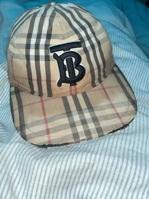 Beige Burberry keps med rutigt mönster - Säljer en klassisk beige keps från Burberry med det ikoniska rutiga mönstret i beige, svart, vitt och rött. Framtill finns ett stort broderat 'TB'-emblem i svart och vitt. Justerbar baktill med tryckknappar. Perfekt för dig som gillar streetstyle och designeraccessoarer.