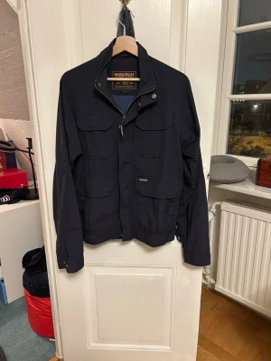 Woolrich fieldjacket  - Stilren mörkblå field jacka från Woolrich med fyra stora fickor framtill, hög krage och dragkedja. Jackan har justerbara muddar och diskret logga på bröstet. Perfekt för blåsiga dagar och enkel att matcha med olika outfits.