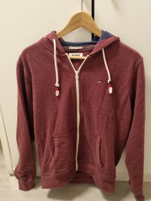 Vinröd hoodie från Tommy Hilfiger - Säljer en vinröd hoodie från Tommy Hilfiger med dragkedja, vita snören och fickor framtill. Liten Hilfiger-logga på bröstet och blått innerfoder i huvan. Perfekt för dig som gillar klassisk streetwear och vill ha något bekvämt och stilrent.