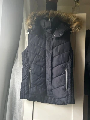 Väst / Gilet - Väst med avtagbar fake päls och avtagbar luva. Endast använd fåtal gånger. Med silver detaljer. Går att använda på olika sätt. Knapparna täcks när man tar bort fakepälsen och även luvan. Med dragkedja och knappar för att hålla värmen. 