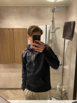 Svart Marcelo burlon hoodie  - Svart Marcelo burlon med huva och färgglada mönster runt halsen och axlarna. Hoodien har en klassisk känguruficka framtill och dragsko i huvan. Perfekt för dig som vill sticka ut med en unik och snygg design.
