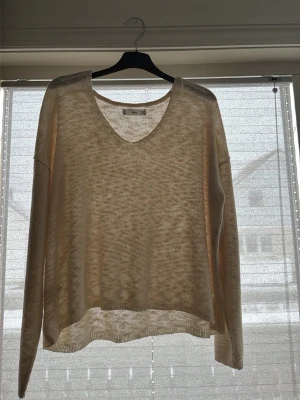 Beige transparent stickad tröja - Säljer en beige, tunnstickad tröja med v-ringning och långa ärmar. Tröjan har en avslappnad passform och är lätt transparent, perfekt för lager på lager. Materialet känns mjukt och luftigt, vilket gör den skön att bära under varmare dagar.