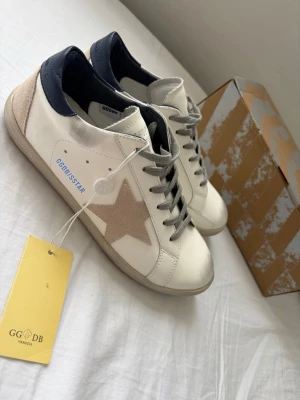 Golden Goose Superstar vita sneakers - ⚠️ÄKTA⚠️Golden Goose Superstar sneakers i vitt läder med beige mockastjärna på sidan, grå skosnören och mörkblå häl. Klassisk låg modell med slitna detaljer och GGDB-tag. Snygg italiensk design som sticker ut.