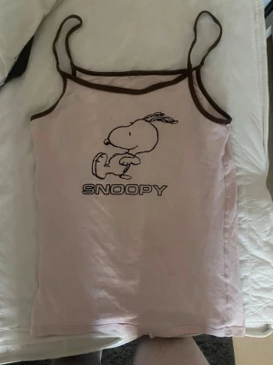 Ljusrosa linne med Snoopy-tryck - Säljer ett ljusrosa linne med tunna axelband och bruna kanter. Framtill finns ett tryck av Snoopy och texten 'SNOOPY'. Perfekt för dig som gillar gulliga och retro motiv. Säljer för det inte är min stil tyvärr xs men passar s 