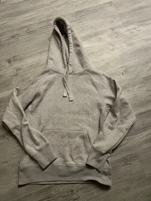 Ljusgrå hoodie med magficka - En stilren ljusgrå hoodie med stor huva och dragsnören. Hoodien har en klassisk magficka framtill och ribbade muddar vid ärmar och nederkant. Perfekt för en avslappnad och casual look. Materialet känns mjukt och bekvämt, troligen bomullsblandning. Tröjan är från russemerch