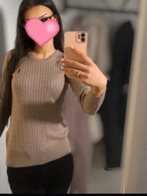 Beige kabelstickad tröja från Ralph Lauren - Snygg beige kabelstickad tröja från Ralph Lauren med rund halsringning och diskret logga på bröstet. Tröjan har en figurnära passform och ribbade muddar vid ärmslut och nederkant. Perfekt för en stilren look.
