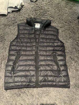 Svart dunväst med dragkedjor - Snygg svart dunväst från McKinley med horisontella sömmar och hög krage. Västen har två framfickor med dragkedja och en diskret reflexdetalj på ryggen. Perfekt för lager-på-lager och enkel att matcha till olika outfits.