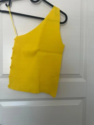 Gul oneshoulder ribbad topp - Snygg gul oneshoulder topp med ribbad struktur och dekorativa knappar längs sidan. Toppen har en smal axelrem och är croppad i modellen, perfekt för en trendig look. Materialet känns mjukt och stretchigt.
