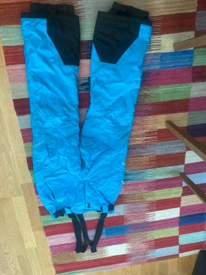 Blå skidbyxor/ täckbyxor McKinley med svarta detaljer strl 40 - Säljer ett par blå skidbyxor från Mc Kinley med svarta förstärkningar vid bensluten och justerbara hängslen. Byxorna har dragkedja och tryckknappar i midjan samt logga på benet. Perfekta för vintersport och utomhusaktiviteter.