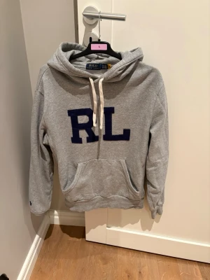 Grå Ralph Lauren Hoodie - Grå hoodie från Polo Ralph Lauren i storlek S. Klassisk modell med huva och dragsnören samt stor mörkblå “RL”-logga framtill. Mjuk och bekväm med magficka framtill – perfekt för en avslappnad men stilren look. Pris går att diskutera 