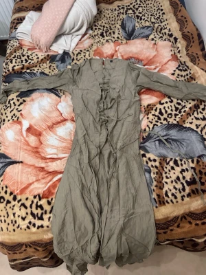 Olivgrön långklänning med snörning - Säljer en olivgrön långklänning med långa ärmar och snygg snörningsdetalj framtill. Klänningen har en avslappnad passform och är tillverkad i ett tunt, lätt material som passar perfekt till varmare dagar. Materialet är limne och behöver därför strykas. Helt oanvänd. 