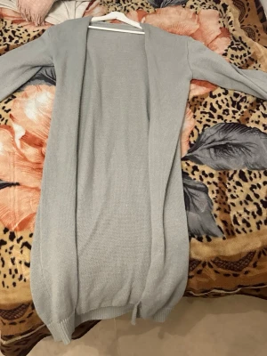 Ljusgrå lång kofta utan knappar - Säljer en stilren ljusgrå lång kofta i mjukt stickat material. Koftan har öppen front utan knappar eller dragkedja, långa ärmar och ribbade muddar vid ärmslut och nederkant. Perfekt att slänga över en t-shirt eller topp för en avslappnad look. Helt oanvänd cardigan. 