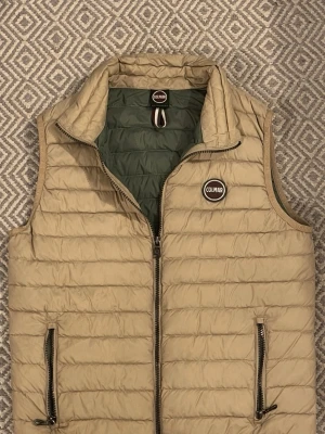 Beige dunväst från Colmar - Stilren beige dunväst från Colmar i storlek XS. Västen har två praktiska fickor med dragkedja och en hög krage. Insidan är olivgrön och märket syns tydligt på bröstet. Väldigt bra skick förutom några mörkare detaljer i botten av framsidan men det går garanterat att få bort. Väldigt snygg nu till våren, kontakta om intresserad eller frågor!!!
