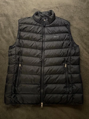 Marinblå dunväst från GANT - Snygg marinblå dunväst från GANT med quiltad design och hög krage. Västen har två dragkedjeförsedda fickor framtill och diskret logga på bröstet. Perfekt att slänga över en hoodie eller tröja för extra värme. Materialet är glansigt och känns riktigt modernt.