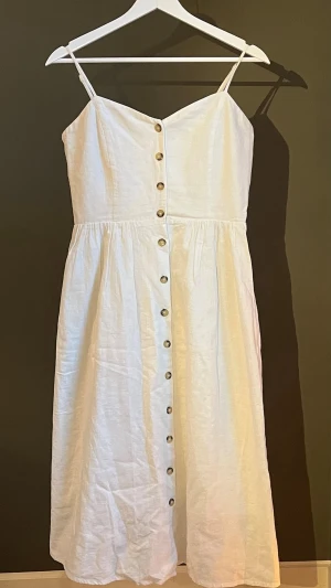 Vit långklänning med knappar - Säljer en vit långklänning med smala axelband och knappar längs hela framsidan. Klänningen har en v-ringad urringning och är tillverkad i ett svalt linneliknande material. Perfekt för varma dagar och enkel att styla med accessoarer. Köpt från H&M🤍