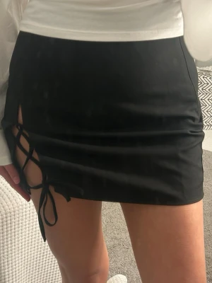 Svart kortkjol med snörning - Snygg svart kortkjol med snörningsdetalj på sidan som ger en edgy look. Kjolen har en tight passform och är perfekt för att skapa en trendig outfit. Tillverkad i ett mjukt och stretchigt material som sitter skönt.