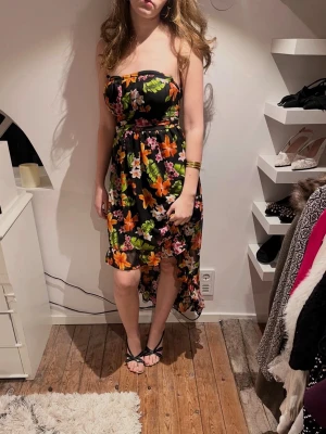 Blommig axelbandslös klänning - Axelbandslös klänning med svart botten och färgglatt blommönster i orange, rosa, grönt och vitt. Klänningen har asymmetrisk längd, kortare fram och längre bak, samt markerad midja. Tunn och luftig, perfekt för varma dagar. Storlek L men passar S.