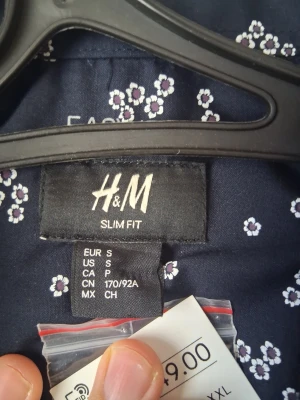 Blommig slim fit skjorta från H&M - Mörkblå slim fit skjorta från H&M med små vita och lila blommor. Skjortan har klassisk krage och knappar framtill. Perfekt för dig som gillar en smalare passform och vill ha en skjorta med lite mer personlighet.