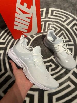 Vita Nike Presto sneakers - Snygga vita Nike Presto sneakers med mesh-ovandel och vita snören. Skorna har en låg silhuett, diskret swoosh-logga på sidan och tå, samt en bekväm och flexibel sula med svart detalj undertill. Perfekta för dig som gillar stilrena och sportiga sneakers.