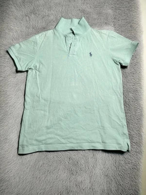 Ljusgrön pikétröja från Ralph Lauren - •  Klassisk Polo Ralph Lauren piké i fräsch mintgrön/turkos – tidlös favorit! 🌿 •  Storlek M – perfekt passform, classic fit med lite rymd (bröst ca 54–55 cm, längd ca 73 cm) •  Äkta detaljer: snygg marinblå ponny-broderad, hög krage, knappar + sidspring •  Skick: Mycket bra! Ren, inga fläckar, inga hål eller noppor – bara normala små brukningsmärken •  Material: Mjuk, skön piquébomull – andas superbra för vår/sommar •  Made in Vietnam – originaletikett finns kvar •  Perfekt till: jeans, chinos.