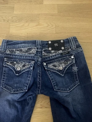 Mörkblå Miss Me jeans - Säljer ett par mörkblå bootcut jeans från Miss Me med snygga slitningar och blingade bakfickor med strass och dekorativa knappar. Jeansen har låg midja. Perfekta för dig som gillar detaljer och vill sticka ut!!❤️❤️