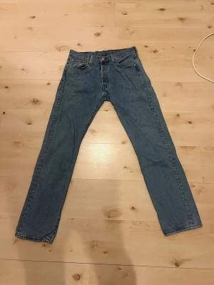 Blå raka jeans från Levi's 501 - Klassiska blå jeans från Levi's med rak passform och fem fickor. Jeansen har en tidlös look med normal midja och är gjorda i slitstark denim. Perfekta för dig som gillar en enkel och clean stil. 