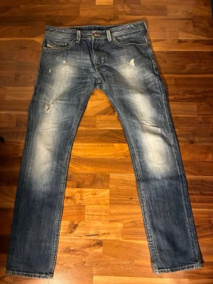 Disel Blå slitna straight jeans - Ett par blå jeans med slitningar och ljusa partier på låren och knäna. Klassisk femficksmodell med raka ben och normal passform. Jeansen har en cool, använd look och är perfekta för en avslappnad stil.