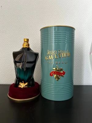 Jean Paul Gaultier Le Beau Le Parfum - Snygg parfymflaska formad som en manlig torso i svart och turkost glas med gulddetaljer. Kommer med en elegant grön metalltub med guldfärgad text och röda detaljer. Perfekt för dig som gillar unika och lyxiga dofter.