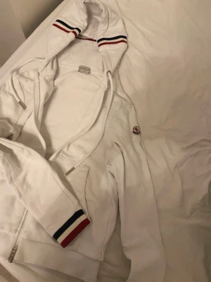 Vit hoodie från Moncler med dragkedja - Säljer en vit hoodie från Moncler med dragkedja framtill och fickor på sidorna. Huva med dragsko och snygga ribbade muddar med röd, vit och marinblå rand. Moncler-logga på ärmen. Perfekt för dig som gillar exklusiv och stilren streetwear.