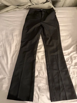 Svarta utsvängda kostymbyxor - Snygga svarta kostymbyxor med utsvängda ben och pressveck. Klassisk midja och diskreta fickor bak. Perfekta för en stilren look och passar till många olika outfits. Materialet känns mjukt och har en lätt glans.