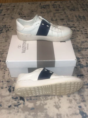 Valentino vita sneakers med blå detalj - Snygga vita sneakers från Valentino Garavani med bred mörkblå läderdetalj över snörningen och vita skosnören. Klassisk låg modell med rund tå och diskret logga på plösen. Tillverkade i slätt läder för en clean och stilren look.