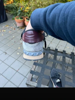 Valentino sneakers med nitar - Snygga sneakers från Valentino med svart mesh och läder, grå mockadetaljer och vinröd häl. Sulan är beige med karaktäristiska pyramidformade nitar i silver på hälen. Skorna har svarta skosnören och en sportig siluett.