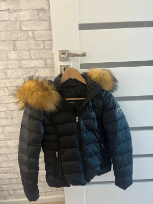 Svart rock and blue jacka  - Snygg svart dunjacka med stor luva och fluffig fuskpäls i gult och brunt. Jackan har quiltad design, dragkedja framtill och två fickor med dragkedja. Märke syns på ärmen. Har även en annan äkta päls till!