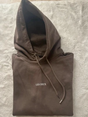 Brun hoodie från Les Deux - Säljer en brun hoodie från Les Deux med vit broderad logga på bröstet. Hoodien har justerbar snörning i huvan och en stor magficka. Tillverkad i mjuk bomull och har en klassisk, normal passform.